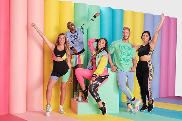 5 adults posing in front of a pastel rainbow wall wearing Zumba Colors of Kindness activewear including shorts, shirts, leggings and jackets. 
